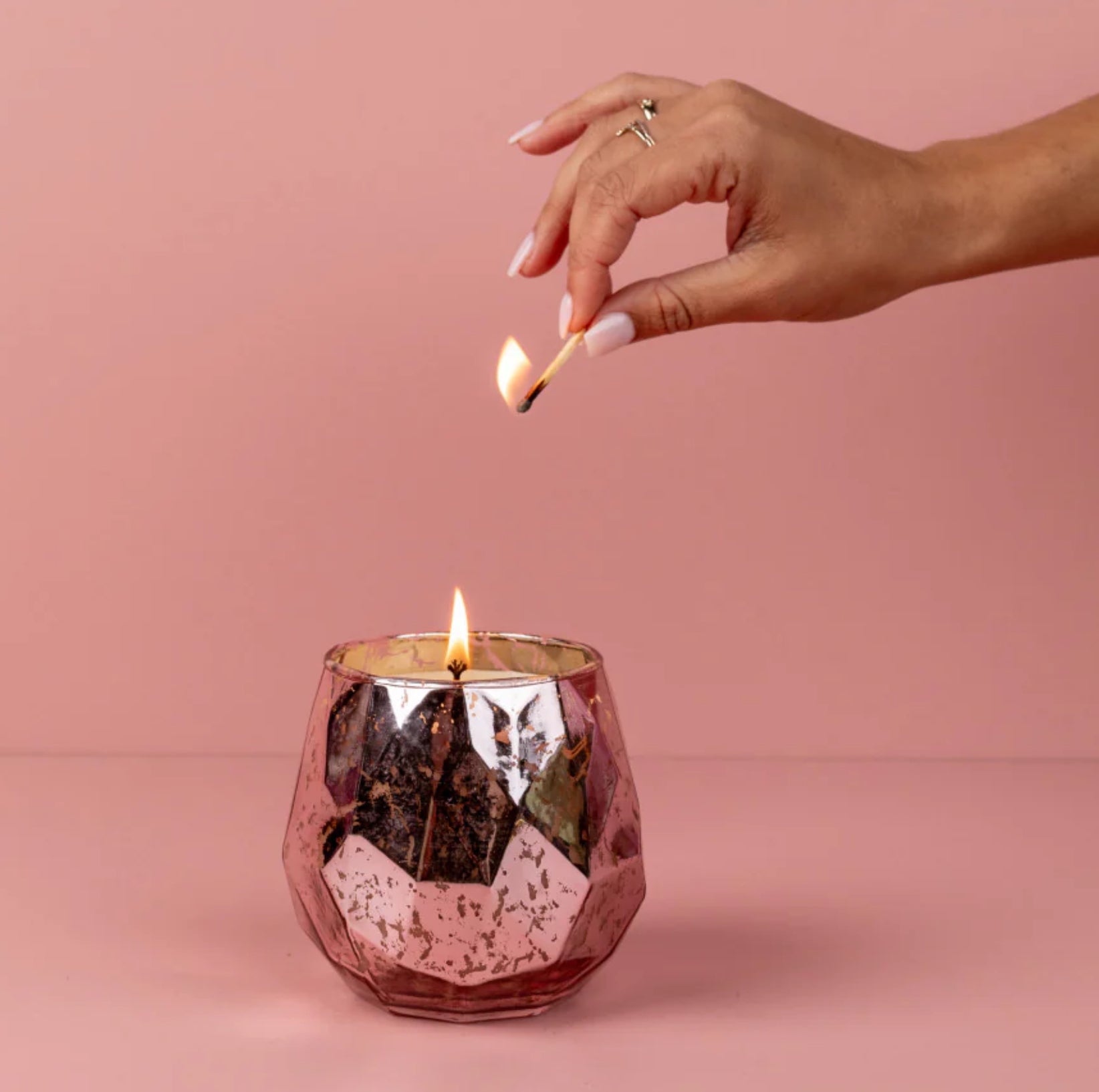 This is a lady lighting a Candle - Sweet Grace - Pink Mercury Glass - 15.8 oz. On a pink background