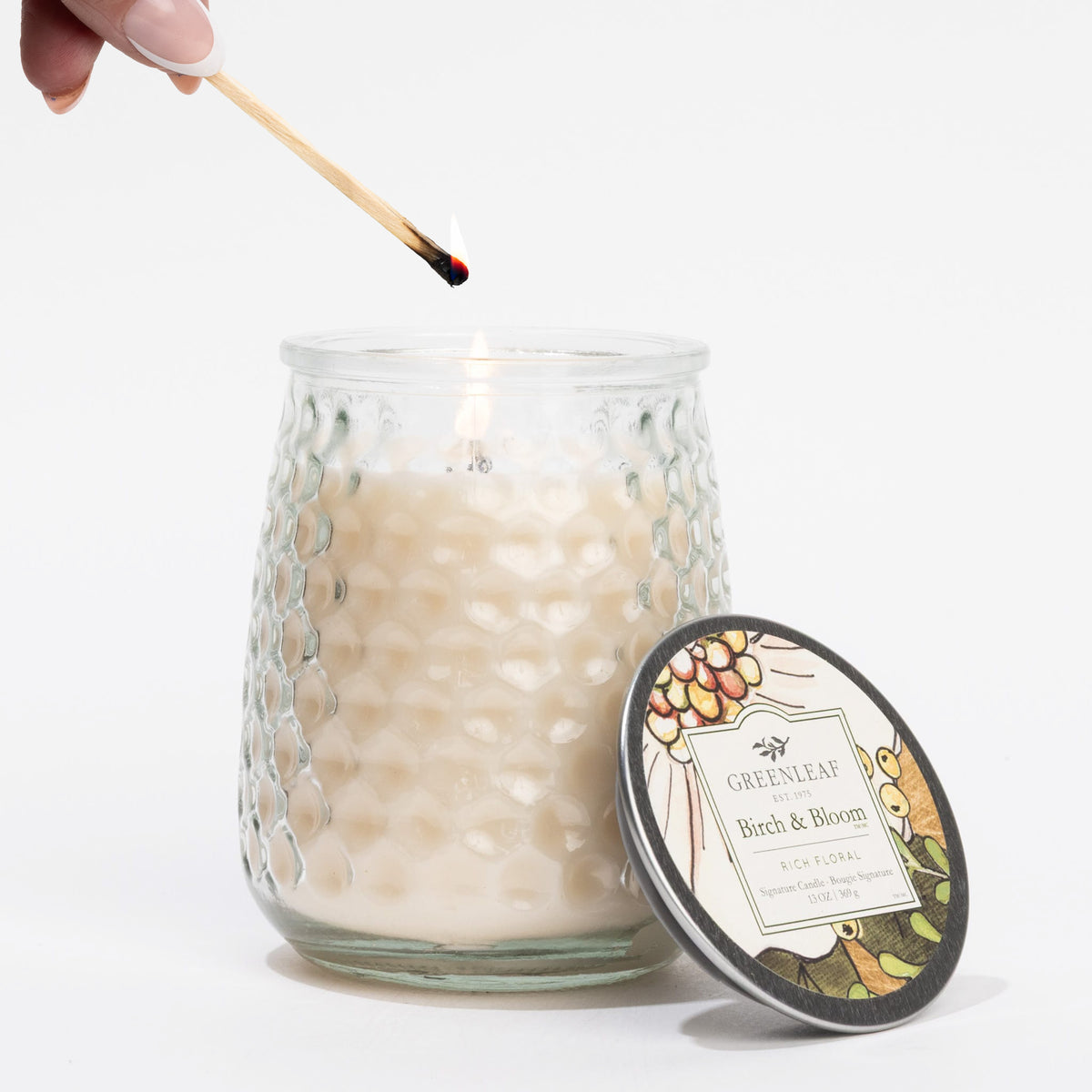 Glass candle with decorative lid labeled 'Greenleaf' being lit with a match.