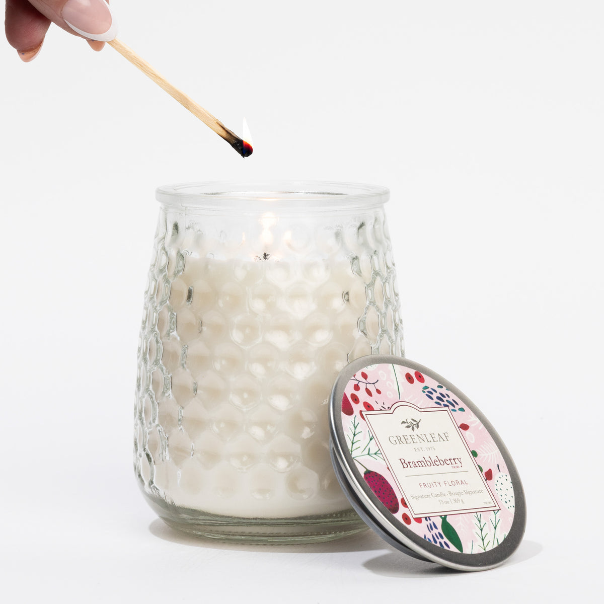 Glass candle with floral label being lit by a match on a white background