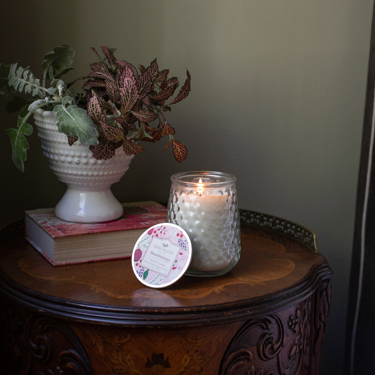 Candle in a textured glass holder on a wooden table with a plant and book in the background.