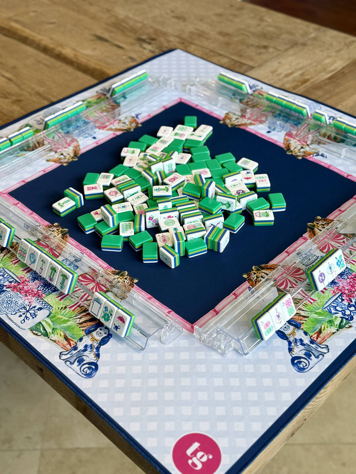A Mahjong game mat  on a wooden table, featuring a white and navy patterned mat, clear acrylic racks, and a pile of green and white tiles in the center.