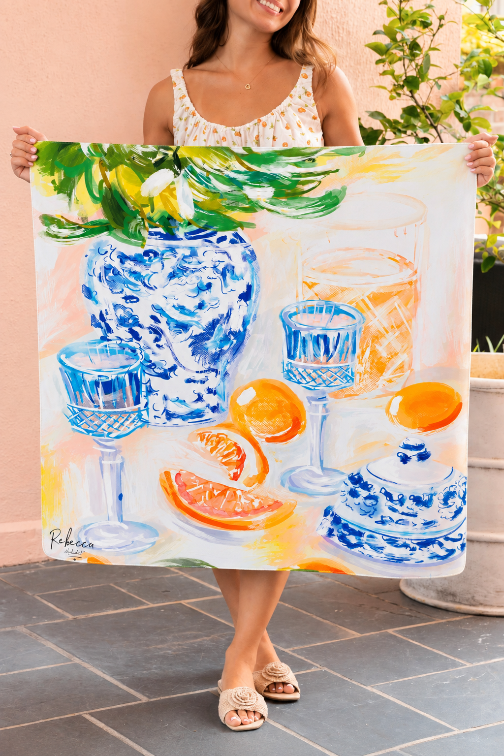 A woman holds up a mahjong mat featuring a colorful still life painting of blue patterned glassware, oranges, and green leaves on a warm cream background.