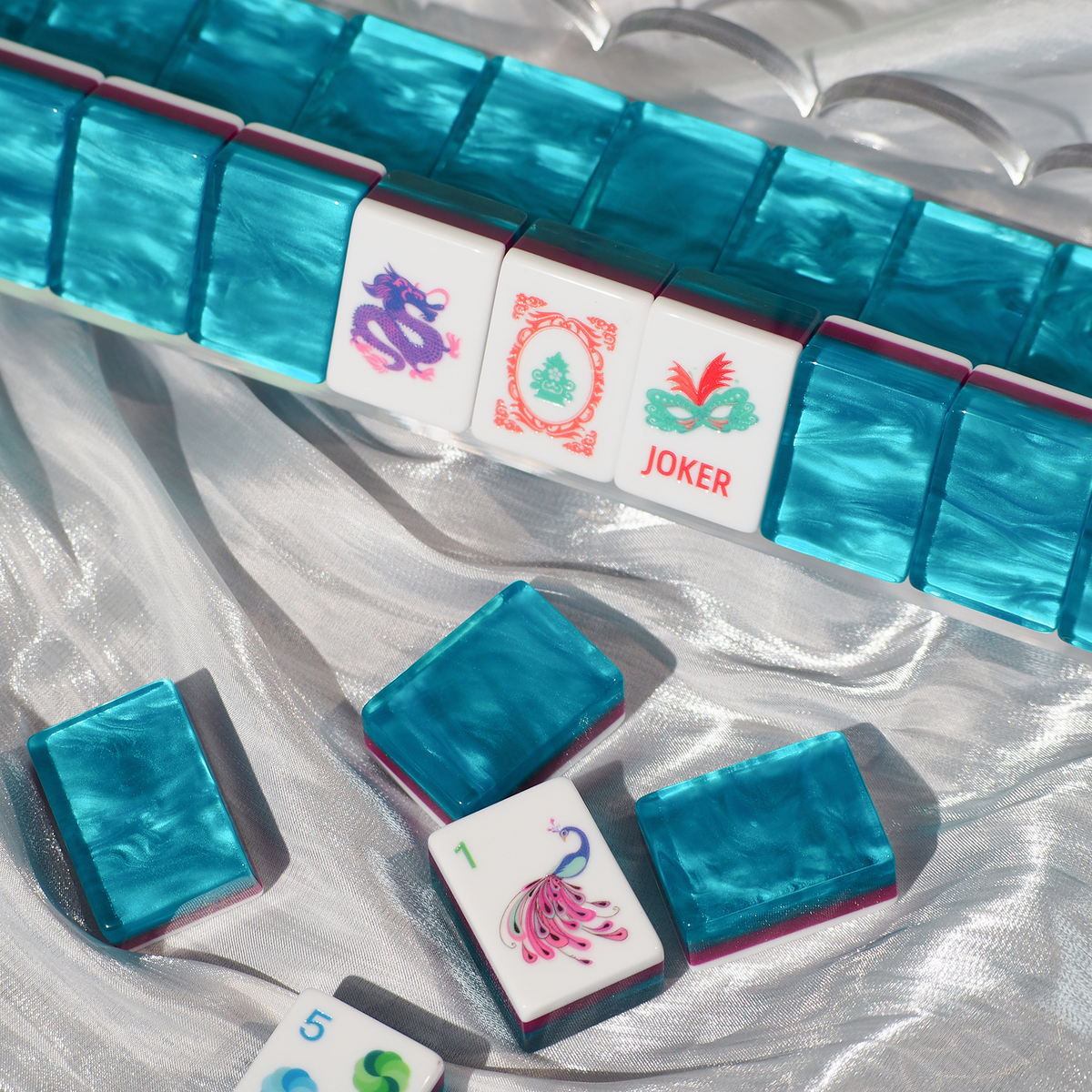 A set of mahjong tiles arranged on a shiny silver fabric background, with several loose tiles in front. The tiles have iridescent turquoise-blue backs and white faces with colorful Chinese characters and symbols, including a joker tile.