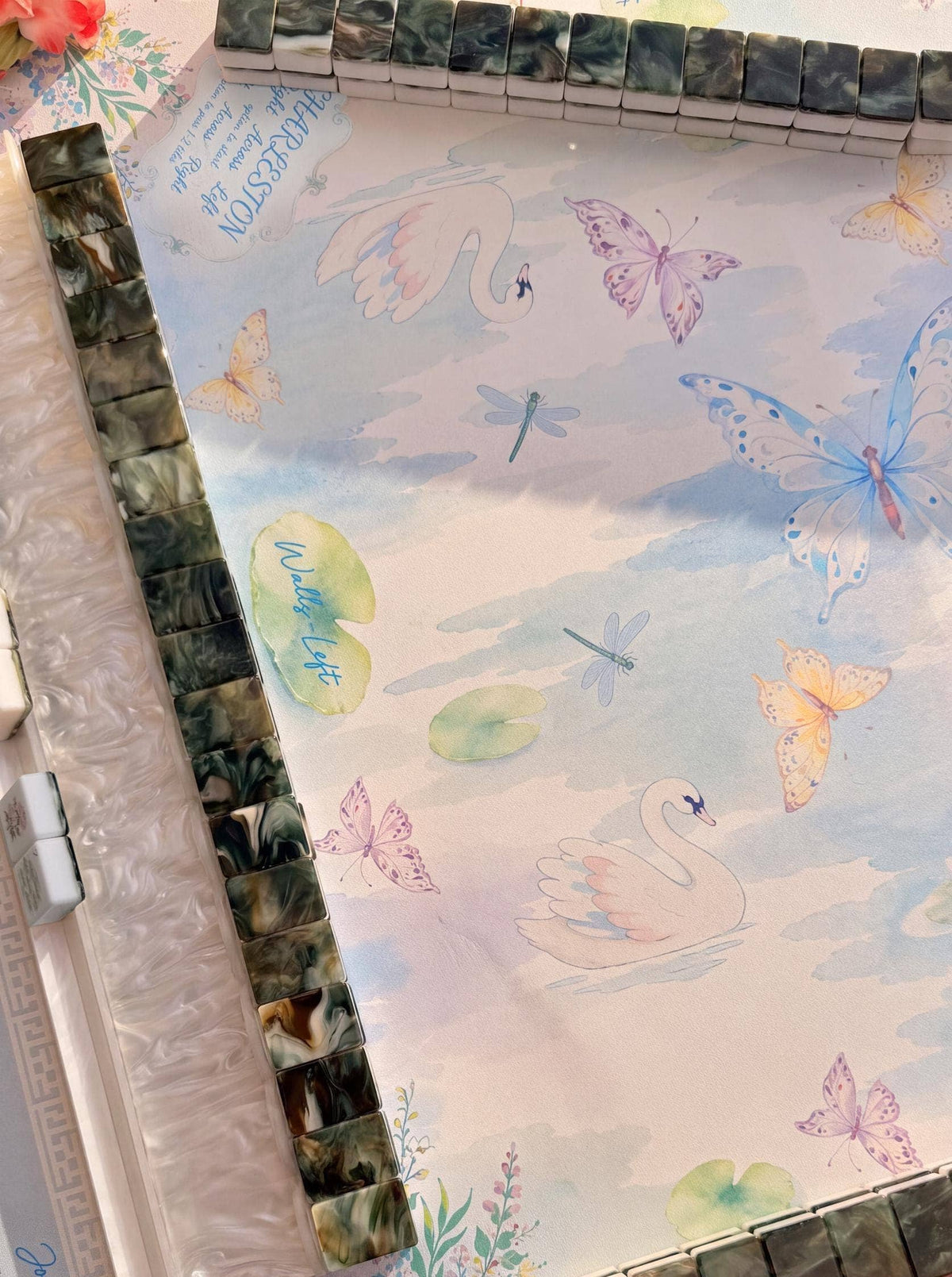 A close-up of a Mahjong Mat featuring a pastel watercolor design with swans, butterflies, dragonflies, and lily pads on a pale background, bordered by glossy marbled tiles.