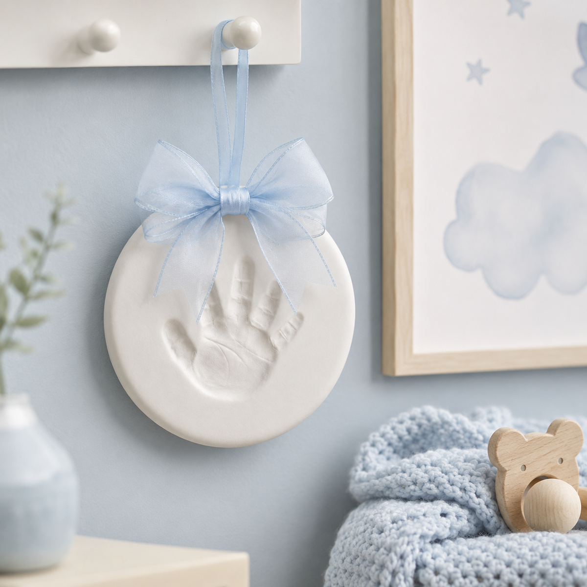 A round white baby keepsake plaque with an embossed handprint hanging from a light blue ribbon bow on a blue wall, styled with a knitted blue blanket, wooden bear pacifier, and framed cloud artwork.