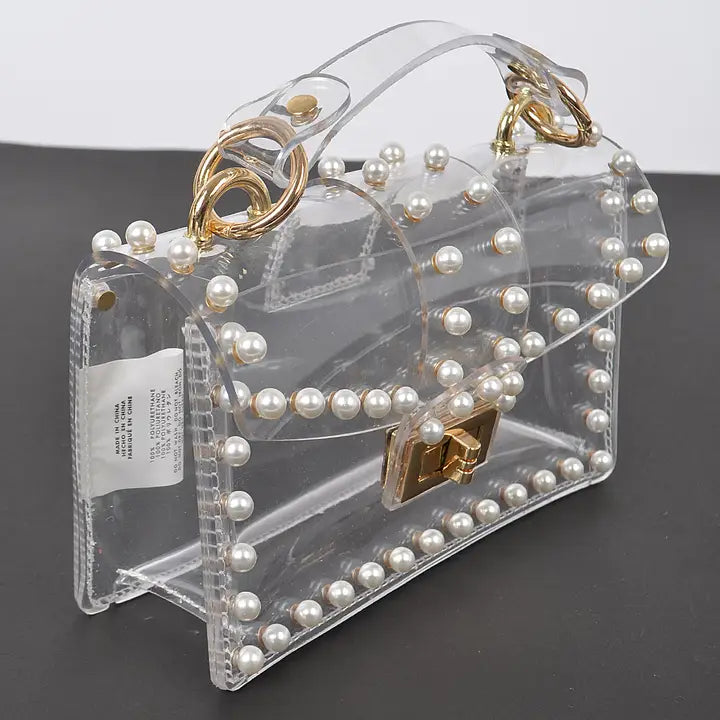 Transparent pearl-studded clear clutch bag with gold twist lock and top handle, angled view on dark background showing full pearl border detail