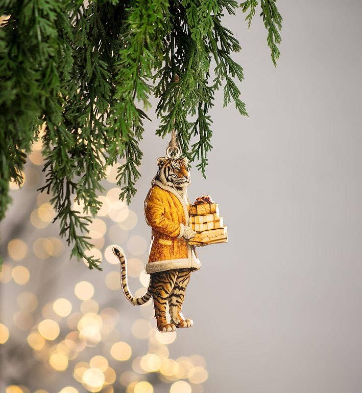 A hanging Christmas ornament shaped like a tiger dressed in a yellow-orange coat and holding wrapped gifts, suspended from green pine branches against a soft blurred background with warm bokeh lights.
