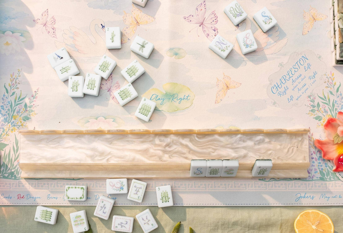 A styled flat-lay of a mahjong game setup featuring white tiles with green and blue floral designs, arranged on a decorative pastel mahjong mat with butterflies and floral illustrations.