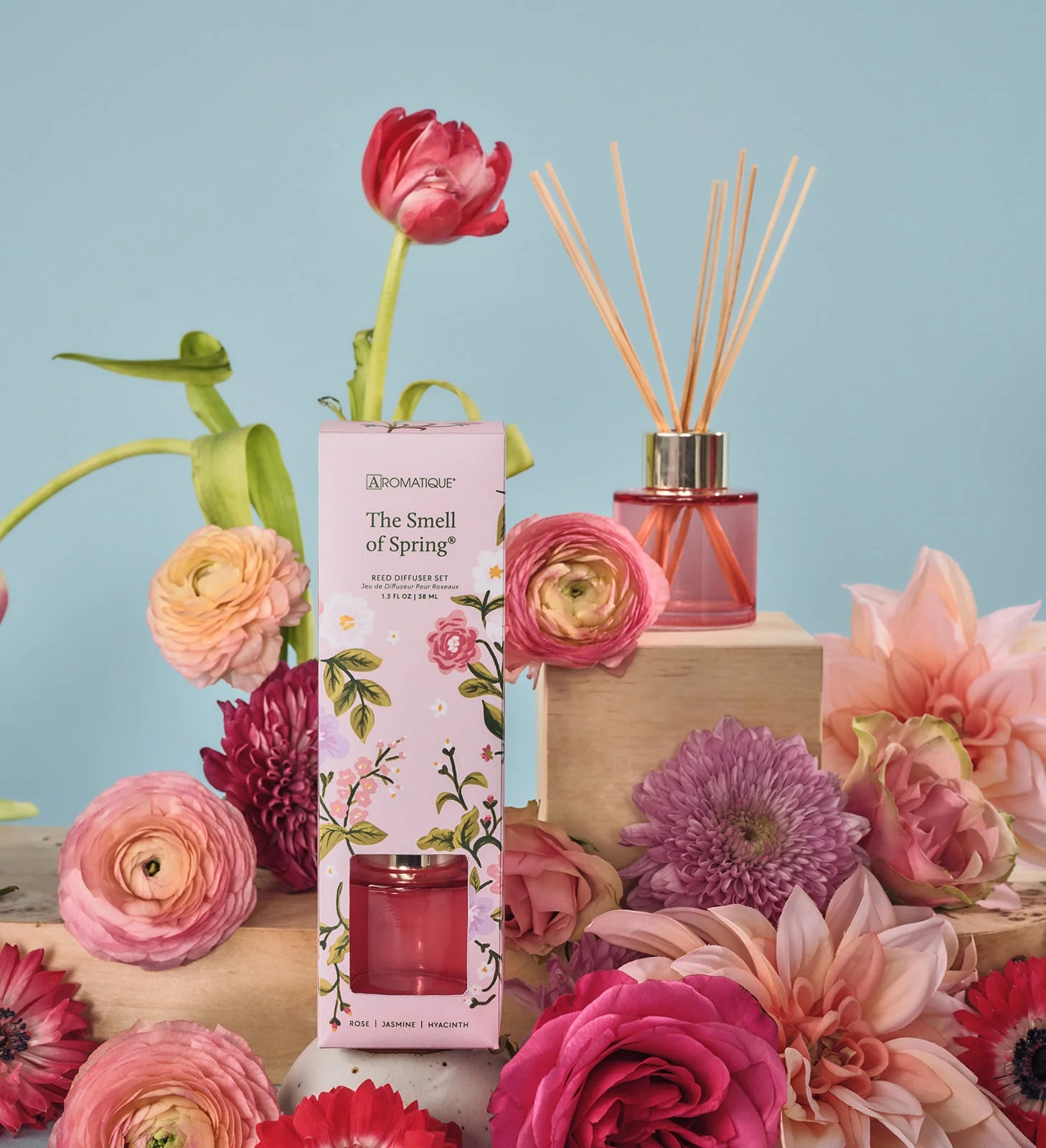 Decorative diffuser and candle with floral packaging surrounded by flowers on a light blue background