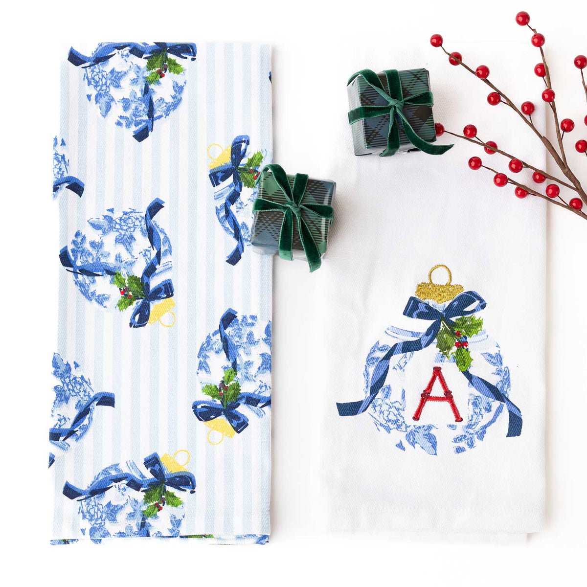 Two white kitchen towels with blue and white holiday patterns are displayed on a white background, styled with two small green gift boxes and red berry branches. One towel features a blue and white toile pattern with blue bows and green holly accents, and the other features a white toile pattern with a red monogram letter 'A' inside a blue ornament design.