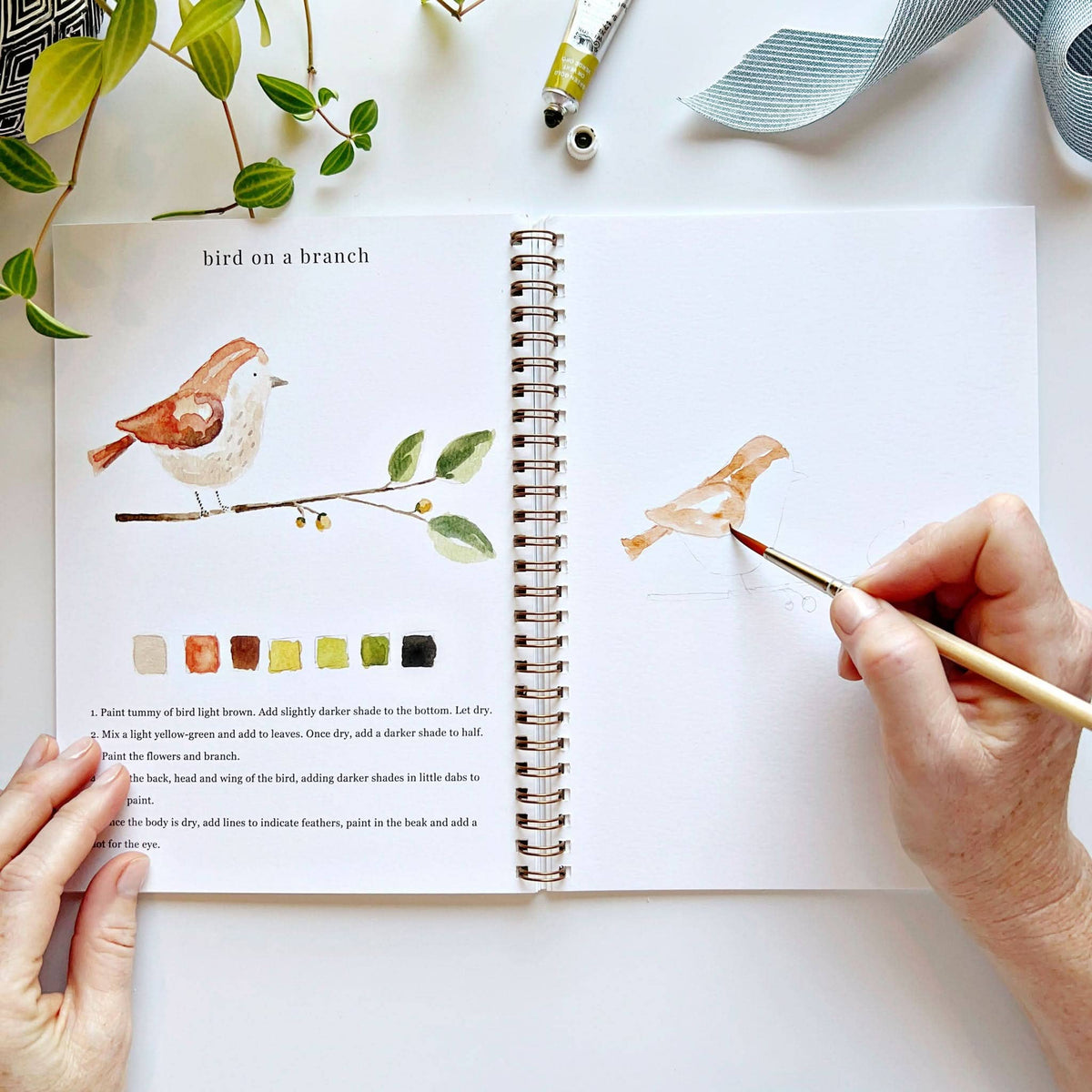 Person painting a bird illustration in a sketchbook with a paintbrush.