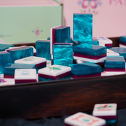 A close-up of stacked and scattered acrylic mahjong tiles in a dark wooden tray, with turquoise, teal, white, and magenta pieces prominently visible against a soft pink and green blurred background.