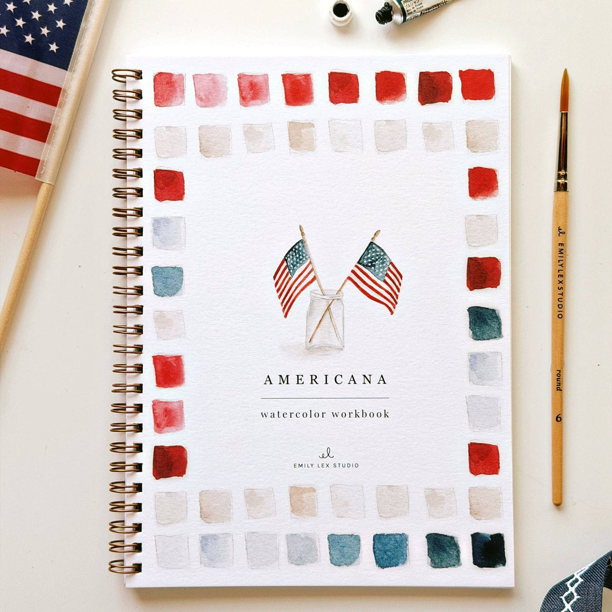 A spiral-bound watercolor workbook titled 'AMERICANA' lies on a white surface, surrounded by a few styled props including a small American flag, paint tubes, and a paintbrush. The cover shows a border of painted color squares in red, white, blue, and gray, with two crossed American flags and the text 'AMERICANA watercolor workbook' and 'EMMY LEX STUDIO'.