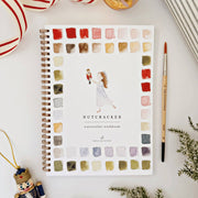 A spiral-bound watercolor workbook titled 'Nutcracker' lies on a white surface, surrounded by a paintbrush, holiday ornaments, ribbon, and greenery. The cover features a watercolor border of color swatches and an illustration of a woman in a white dress holding a nutcracker figure.
