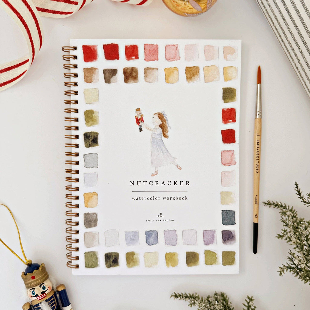 A spiral-bound watercolor workbook titled 'Nutcracker' lies on a white surface, surrounded by a paintbrush, holiday ornaments, ribbon, and greenery. The cover features a watercolor border of color swatches and an illustration of a woman in a white dress holding a nutcracker figure.