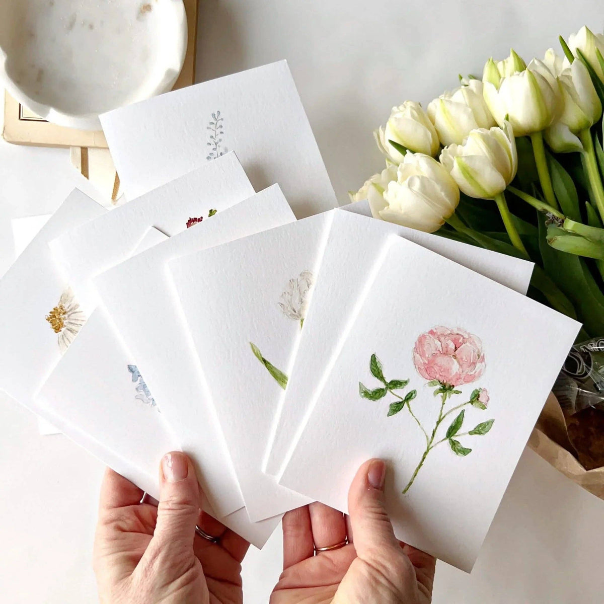 Set of floral cards held by hands with a bouquet of white tulips in the background.