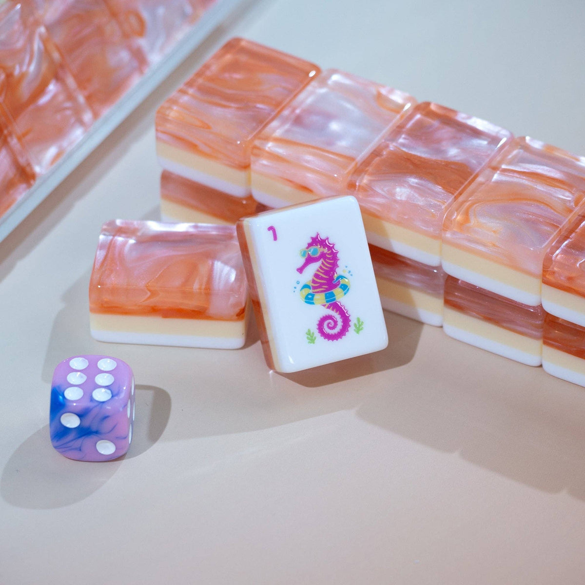 A set of marbled acrylic mahjong tiles with peach, orange, and cream layers, alongside a purple and blue marbled dice, arranged on a light beige surface.