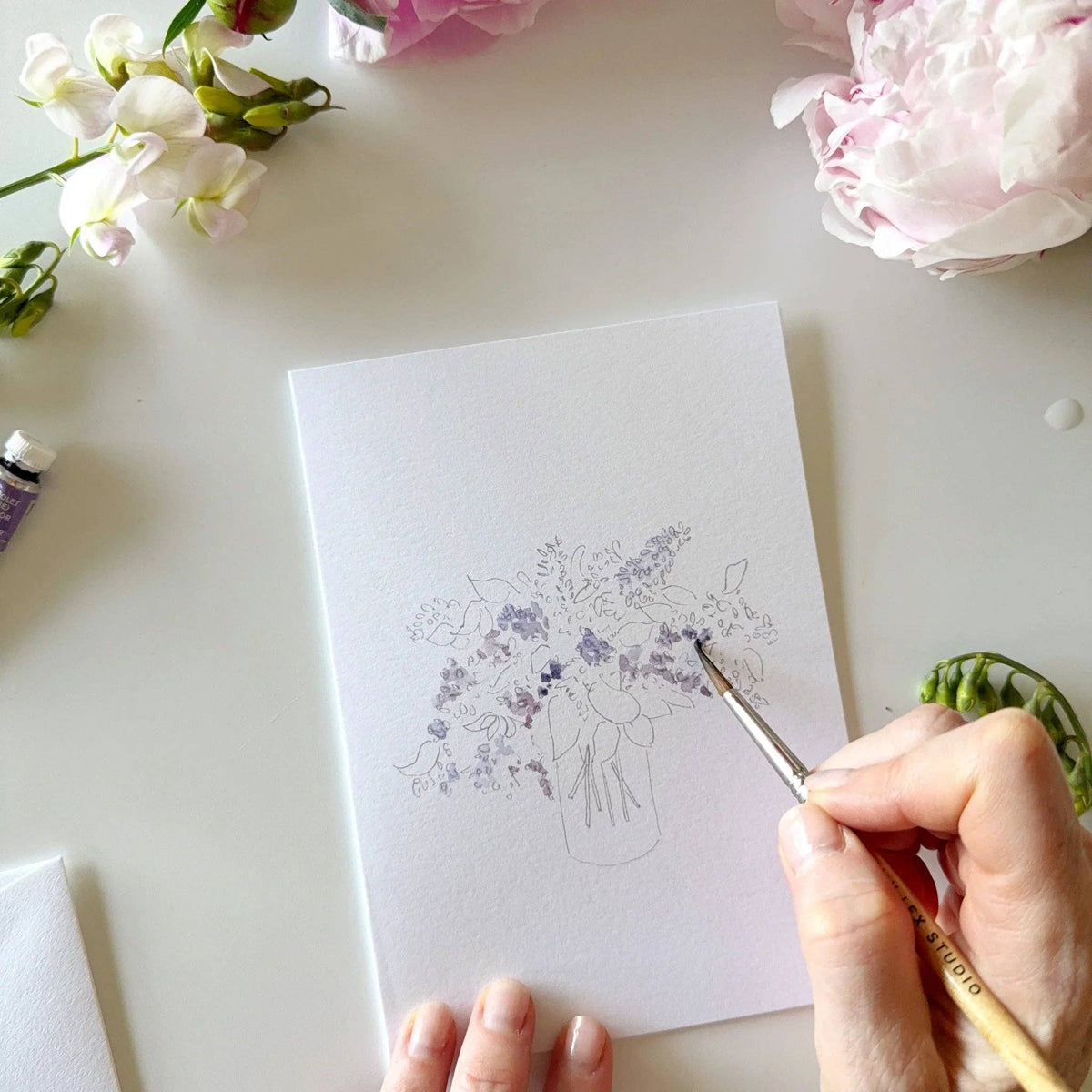 Person drawing a floral design on paper with flowers and art supplies around