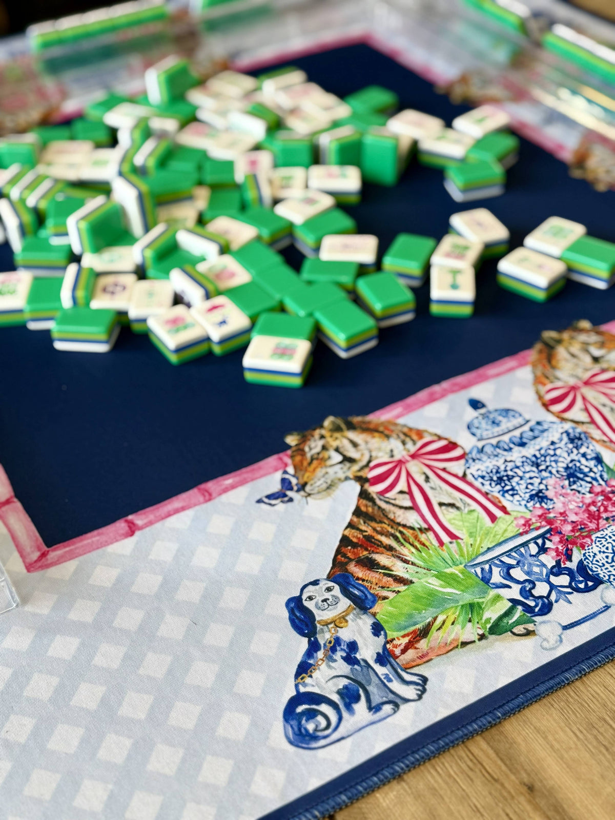 A close-up of a decorative mahjong mat with green and white tiles scattered across the navy blue mat. The mat features a colorful printed border with a tiger, a blue-and-white dog, and a blue-and-white checkered section, chinoiserie jar photographed on a light wood surface.