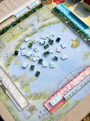 A Bloomjong brand Mahjong game set laid out on a decorative printed mat featuring a garden scene. The mat shows a light blue sky and floral landscape border, with several green-backed tiles and white tiles with colored characters arranged in the center.
