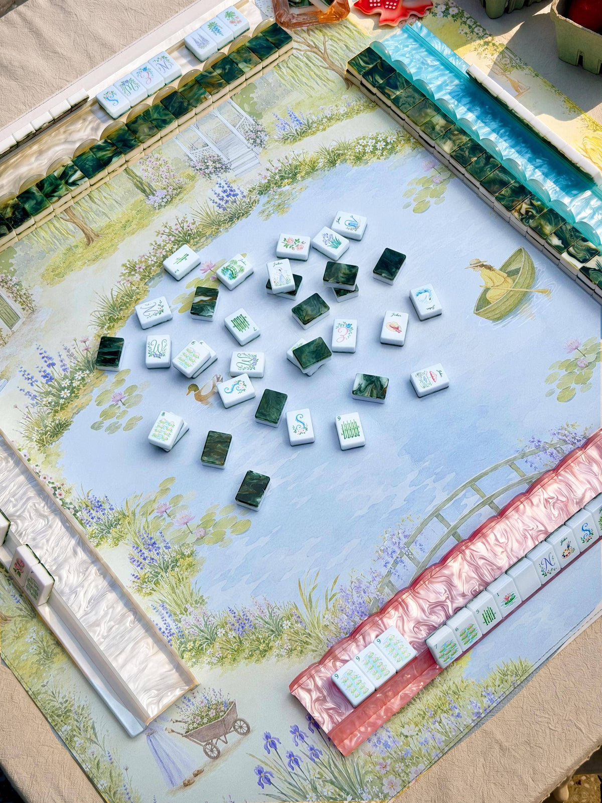 A Bloomjong brand Mahjong game set laid out on a decorative printed mat featuring a garden scene. The mat shows a light blue sky and floral landscape border, with several green-backed tiles and white tiles with colored characters arranged in the center.