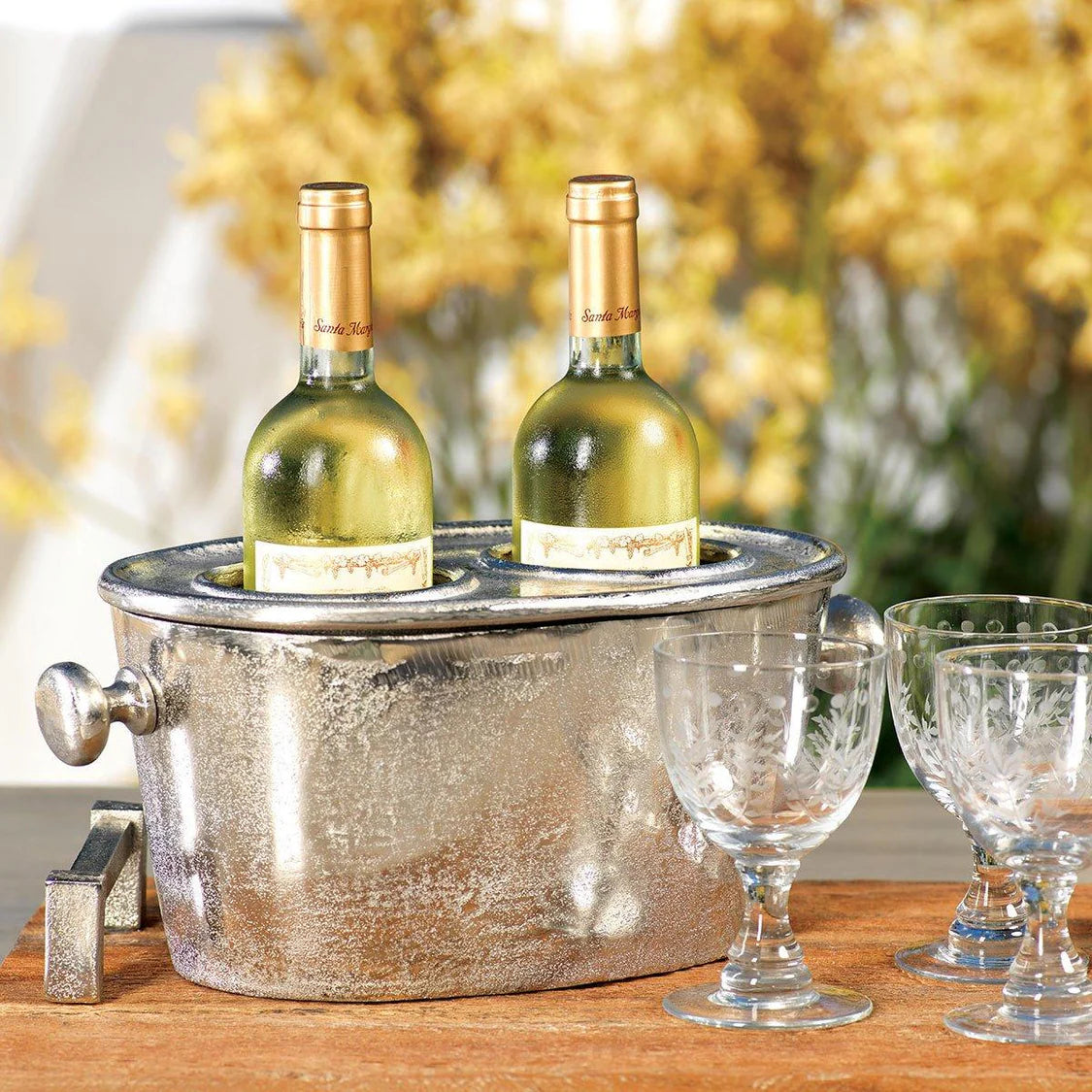 Two wine bottles in a metal ice bucket with glasses on a wooden surface.