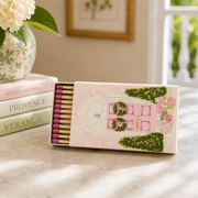 A decorative matchbox with a pink door illustration and greenery sits open on a marble surface beside stacked books and a vase of flowers in a softly lit interior.