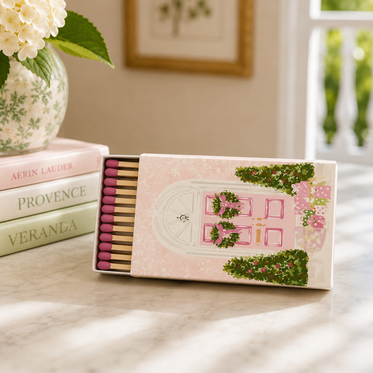 A decorative matchbox with a pink door illustration and greenery sits open on a marble surface beside stacked books and a vase of flowers in a softly lit interior.
