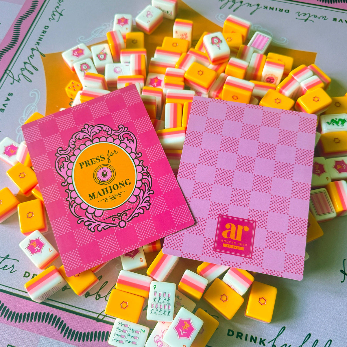Two pink branded shufflers cards and  mahjong yellow, orange and white tiles  on a decorative mat. The visible text/logo on the boxes reads AR and Press for Mahjong.