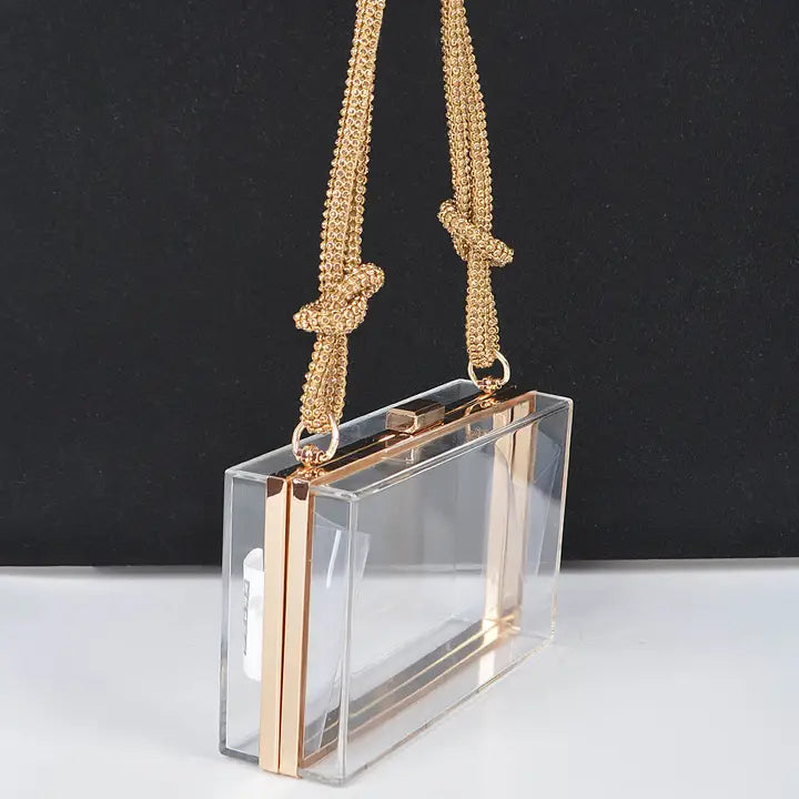 Clear acrylic case clutch with gold metal frame and multi-strand knotted gold chain strap, side angle view on dark background