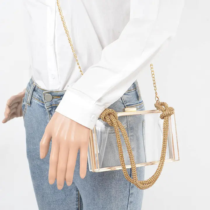Woman in white blouse and jeans carrying clear acrylic box clutch as a crossbody with gold chain strap, lifestyle view showing wearable size