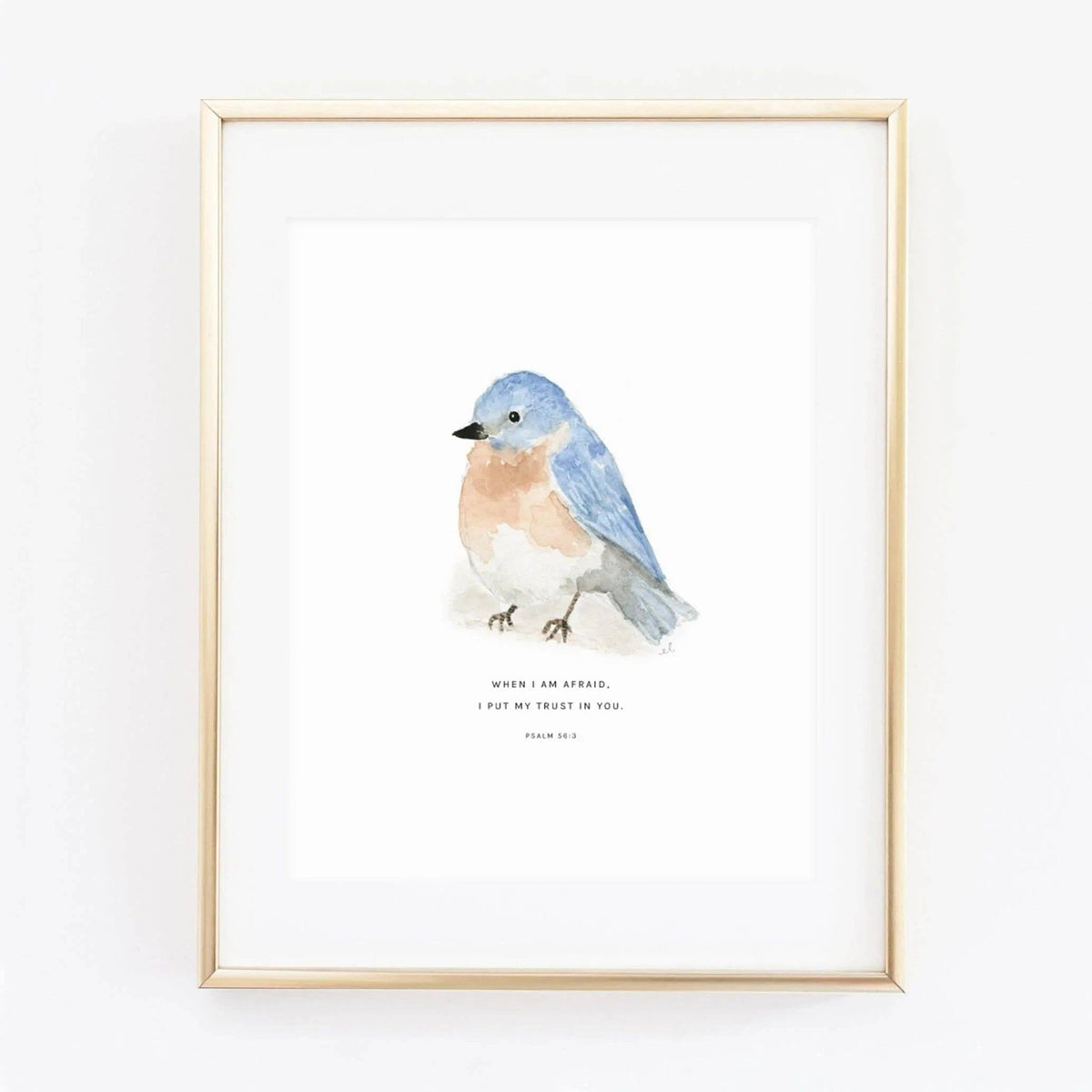 Framed wall art featuring a watercolor bluebird on a white background with a small printed quote beneath it. The piece is shown in a thin light gold frame against a clean white wall.