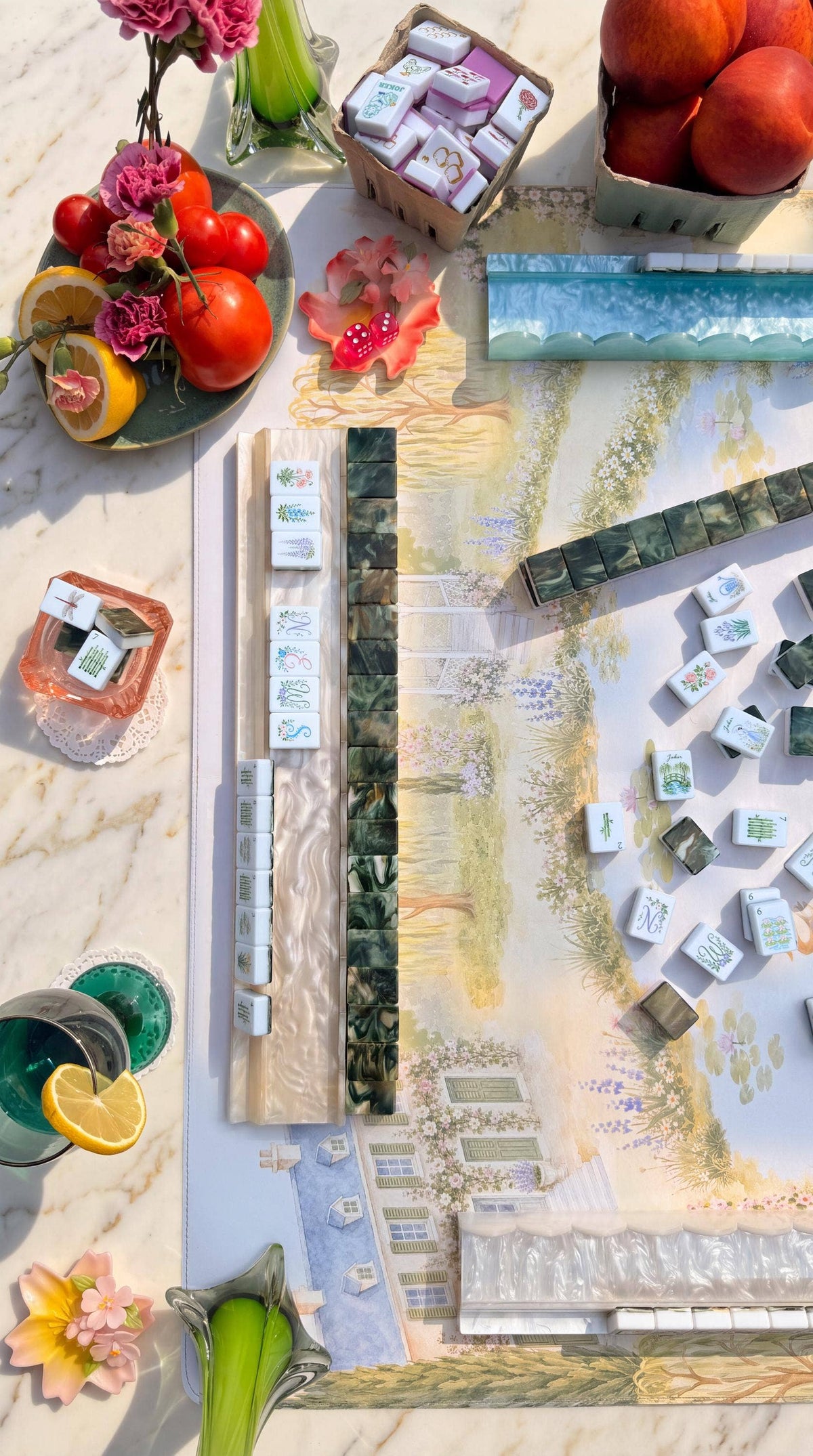 A styled overhead flat-lay of a mahjong game set on a marble surface, surrounded by decorative props including tomatoes, flowers, fruit, and drinks. The set includes a printed board with illustrated tiles, several racks, and many small rectangular tiles with visible brand markings.