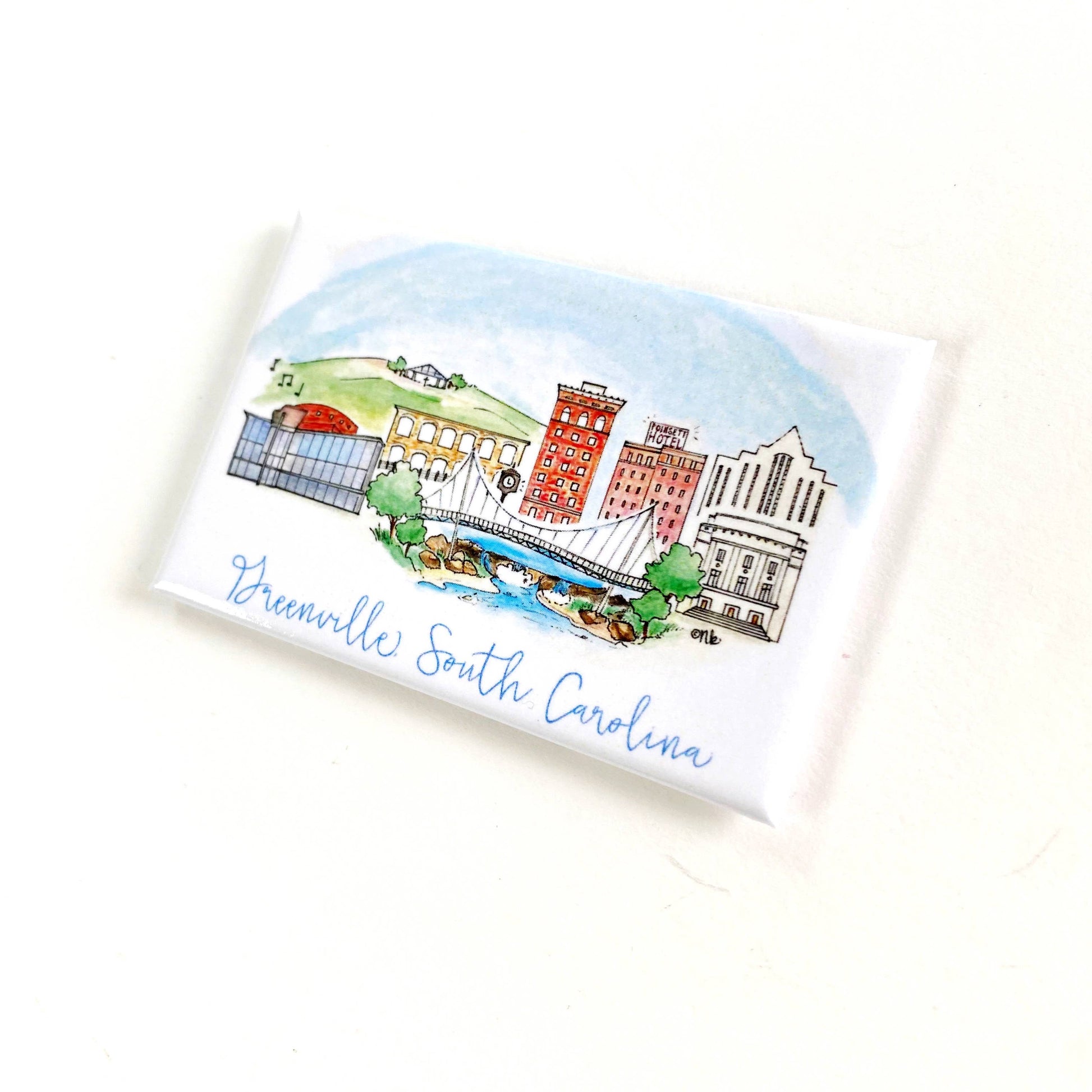 A small rectangular white card or coaster featuring a hand-drawn illustration of Greenville, South Carolina, with blue script text at the bottom, photographed on a plain white background.