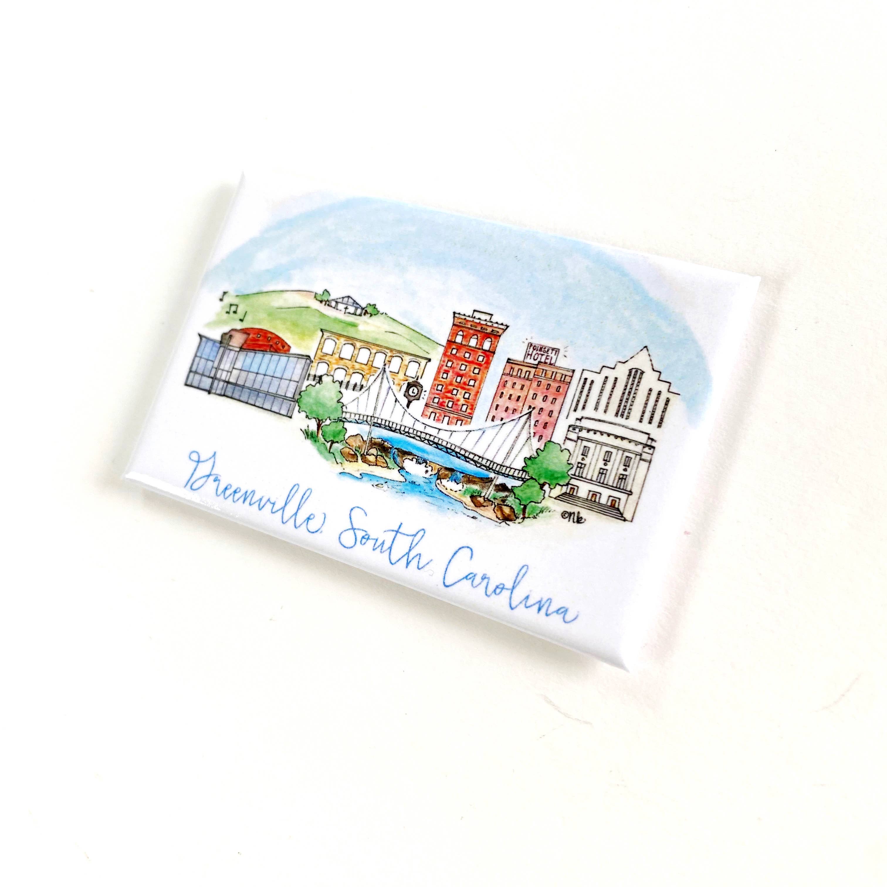 A small rectangular white card or coaster featuring a hand-drawn illustration of Greenville, South Carolina, with blue script text at the bottom, photographed on a plain white background.