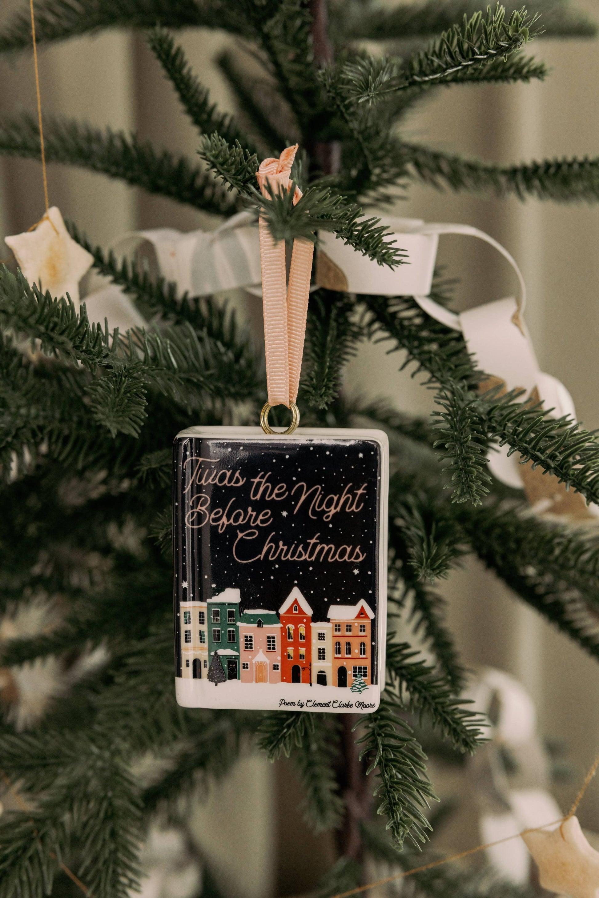 A Steel Mill brand rectangle Christmas ornament that is nceramic book shape hanging from a green tree branch, featuring the phrase 'Twas the Night Before Christmas' in white script over a dark starry background, with illustrated colorful houses along the bottom.