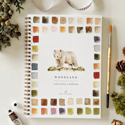 Notebook with bear illustration and 'Woodland Watercolor Workbook' text on a white surface with decorative elements.