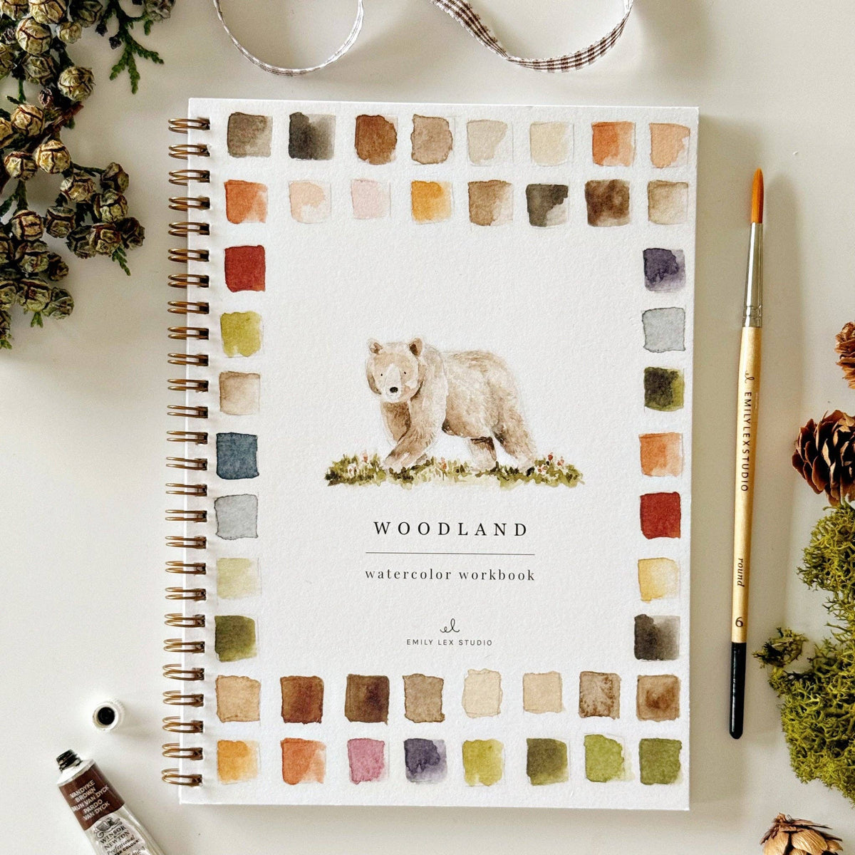 Notebook with bear illustration and 'Woodland Watercolor Workbook' text on a white surface with decorative elements.