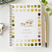 Garden watercolor workbook with color swatches and plants on a white surface with art supplies.
