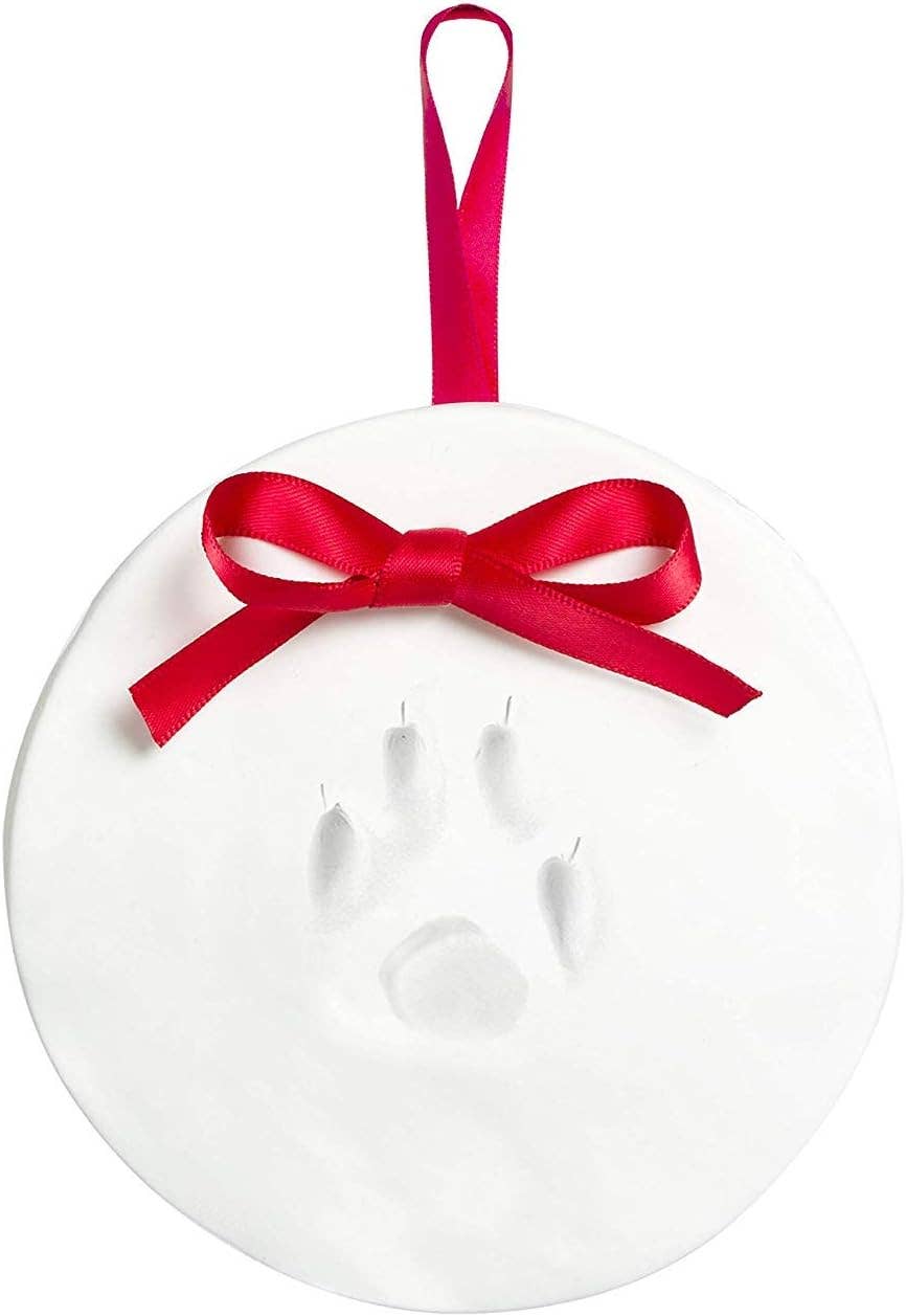 A round white ornament featuring a paw print imprint in the center, decorated with a bright red satin bow and a matching red hanging ribbon, shown on a plain white background.
