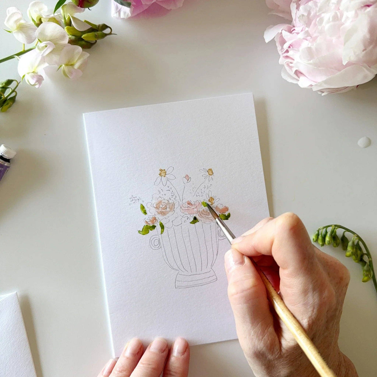 Person painting a floral design on a card with flowers and art supplies around
