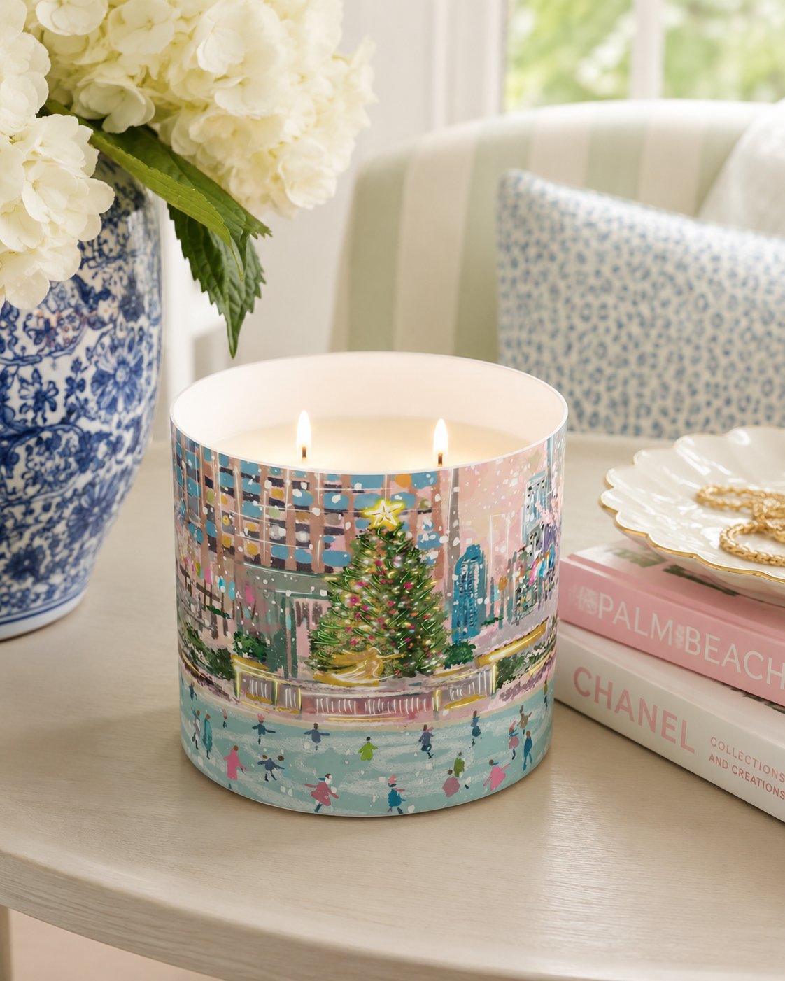 A double-wick candle in a cylindrical container with a colorful winter city scene printed on the outside, lit with two flames. The candle sits on a light wood table next to a blue-and-white ceramic vase with white flowers, a stack of pink and white books, and a decorative dish with gold jewelry in a softly lit interior setting.
