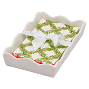 A white ceramic gift tray with a scalloped edge, holding a guest/dinner size paper napkins wiith a green holly wreath pattern and tied with a white ribbon bow, on a plain white background