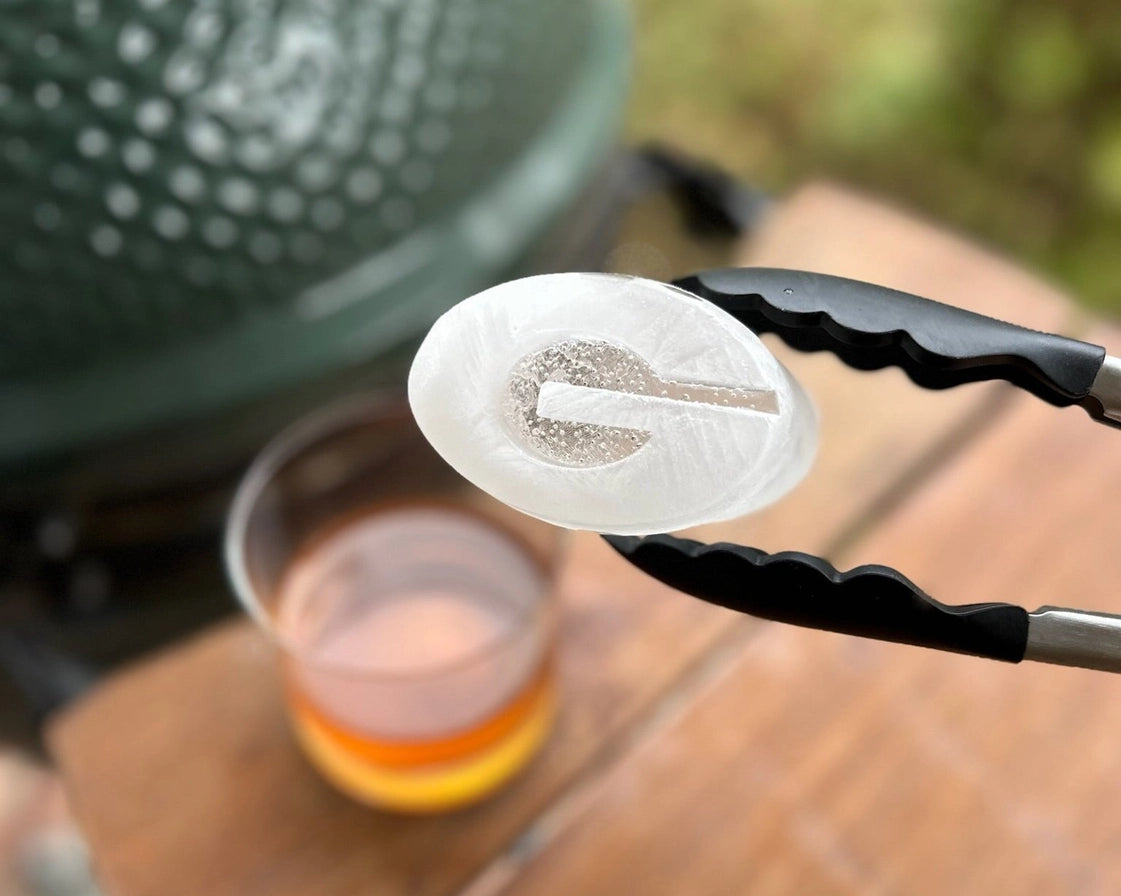 Tongs holding a round white object with a logo over a grill and cup on a wooden surface
