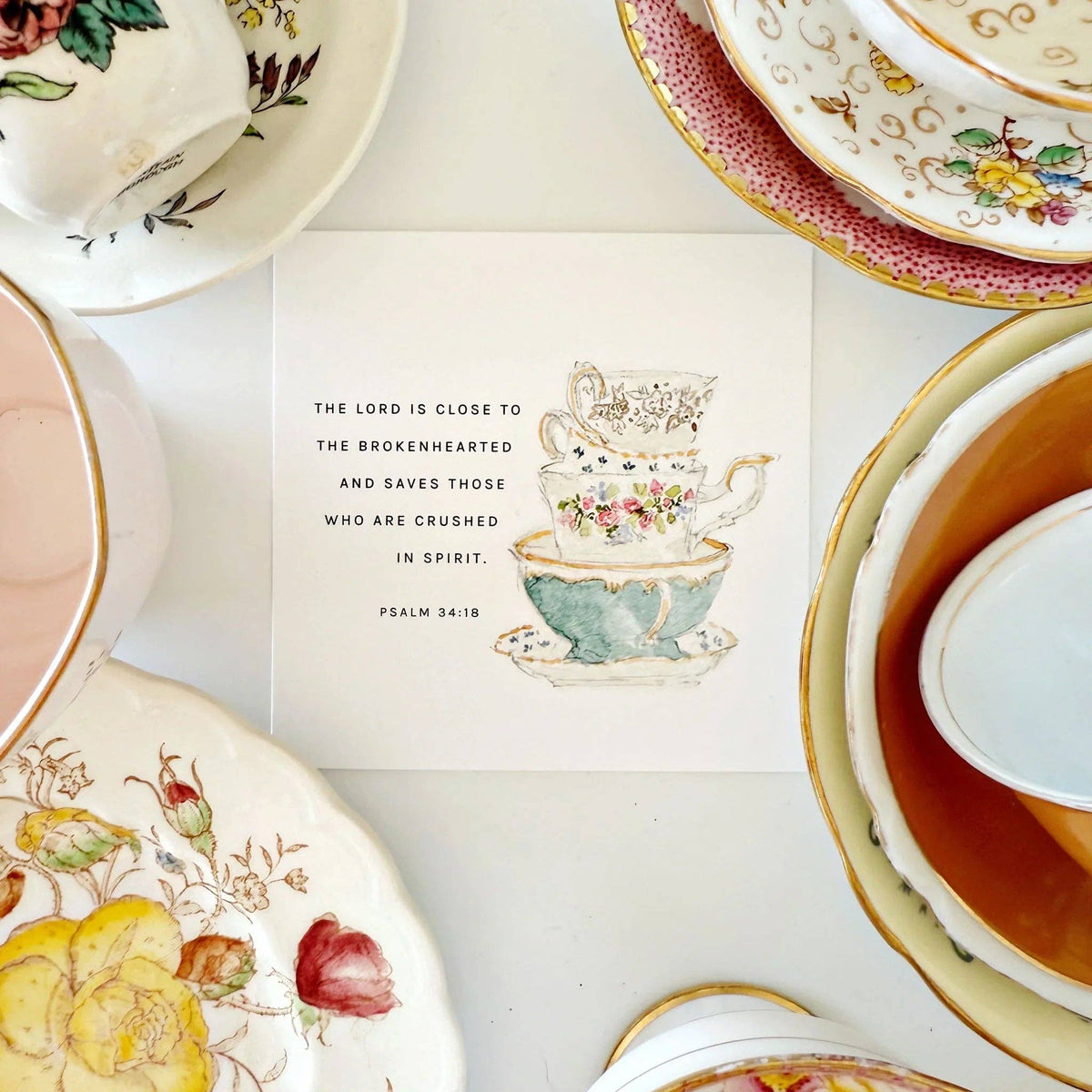 Decorative teacups and plates with a religious quote card in the center on a white background.