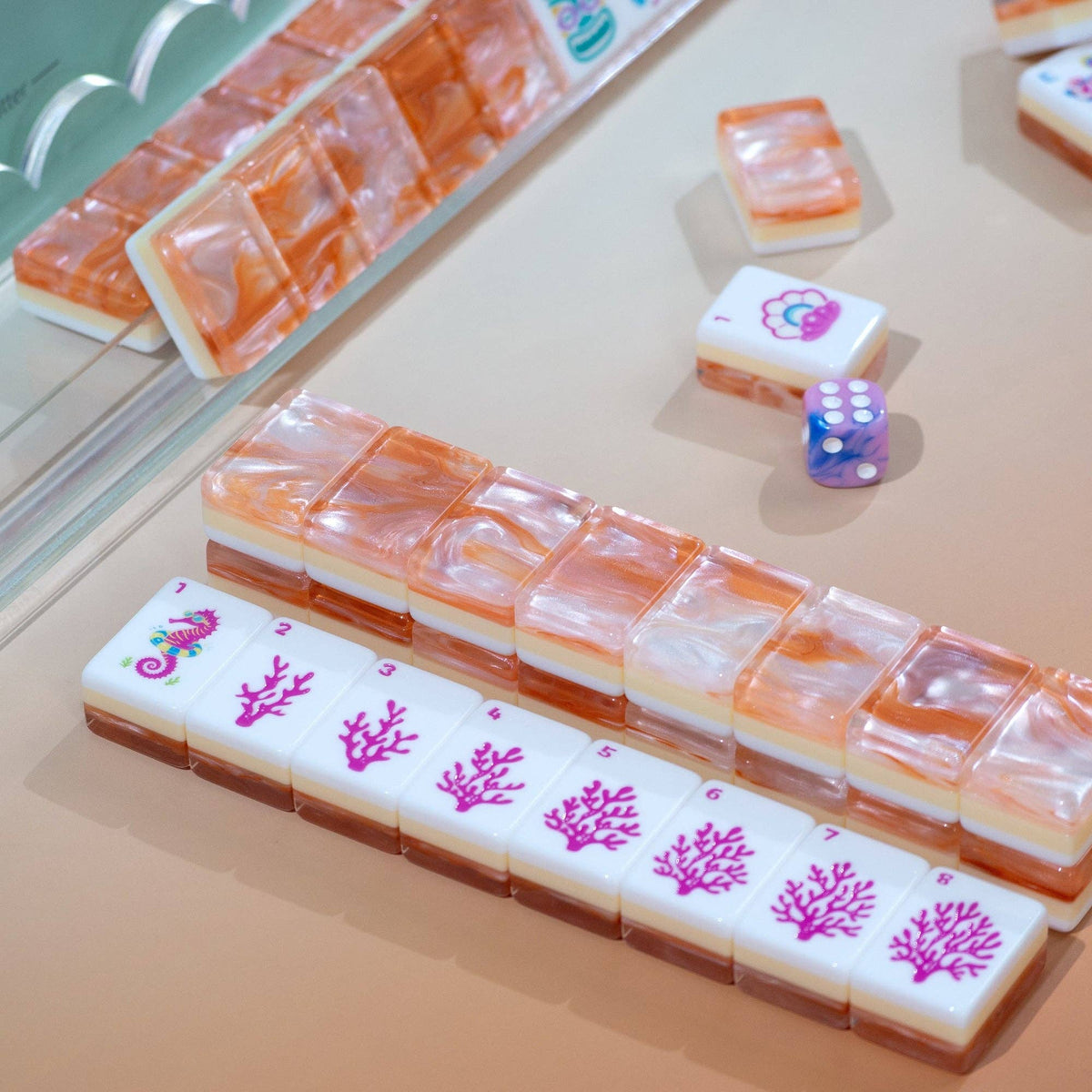 Mahjong-style tiles arranged on a beige tabletop, with translucent orange-and-white layered tiles and white tiles printed with pink coral motifs. A small purple die is also visible.
