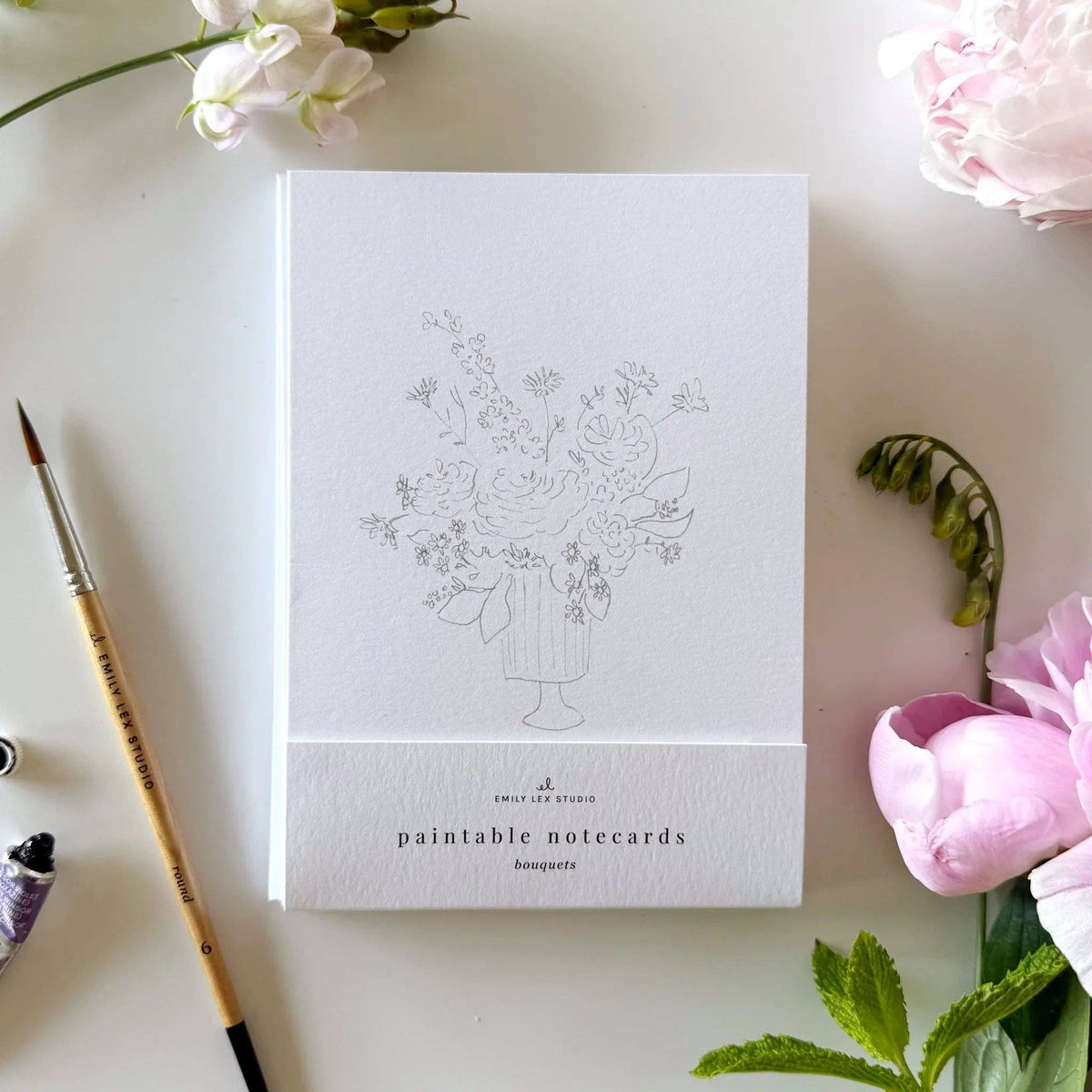 Paintable notecard with floral design on a white surface with flowers and a paintbrush.