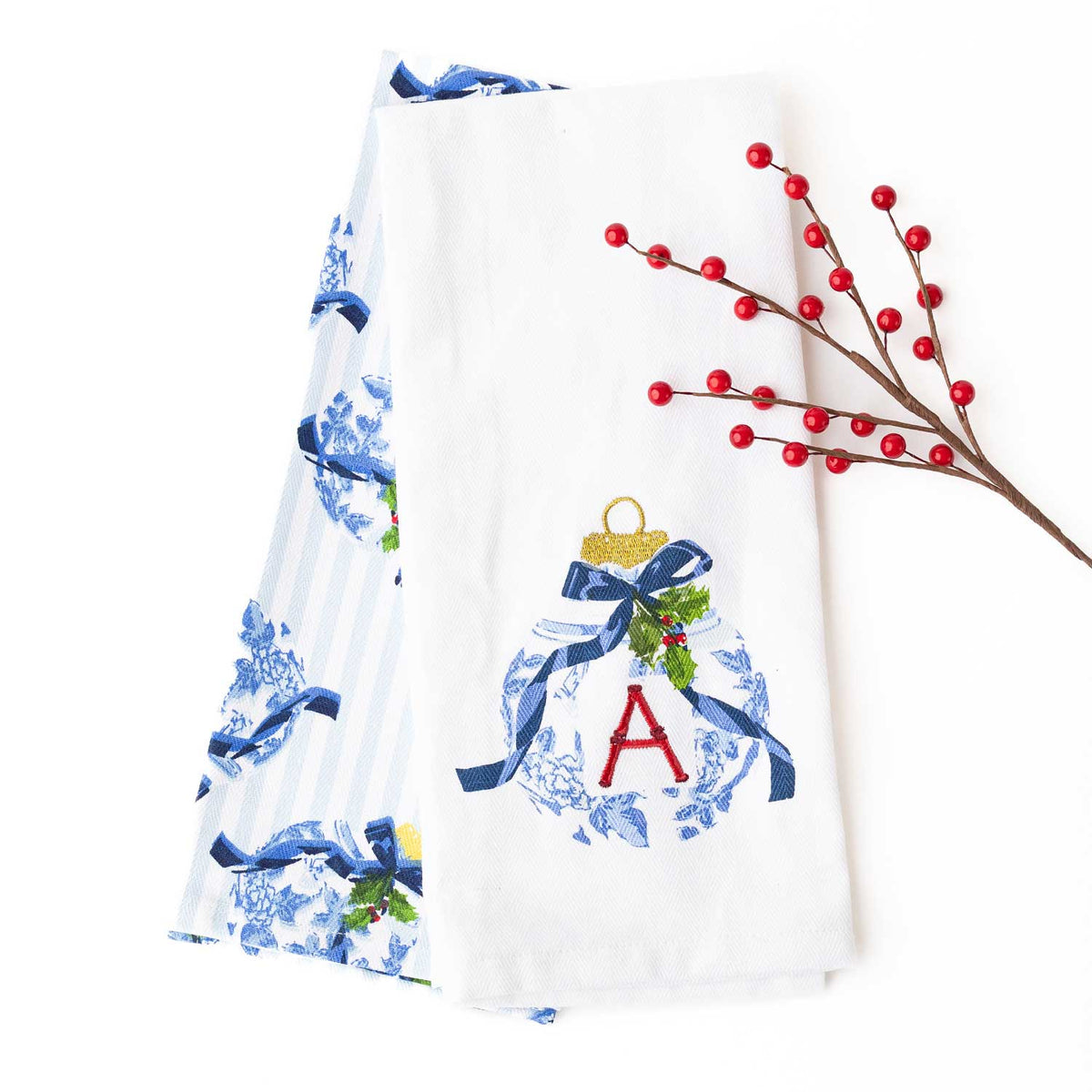 A white kitchen towel with blue floral and striped patterns, featuring an embroidered Christmas ornament design with a red letter 'A' and a decorative branch with red berries on a white background.