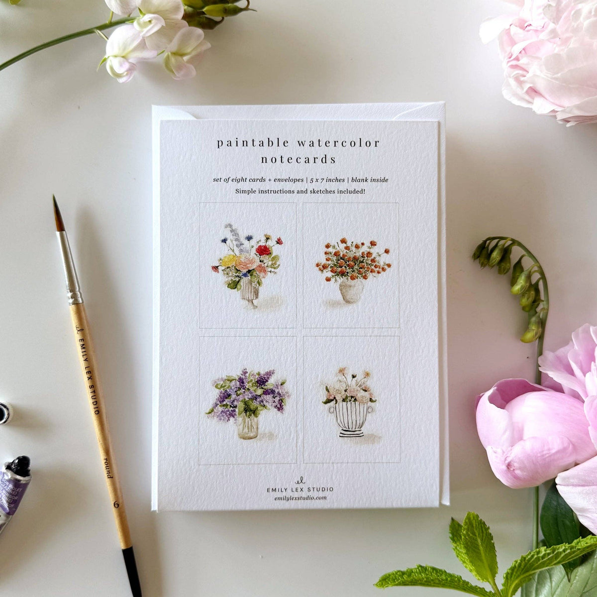 A boxed set of paintable watercolor notecards by Emily Lee Studio, styled in a flat lay with a watercolor brush, flowers, and green leaves on a light surface.