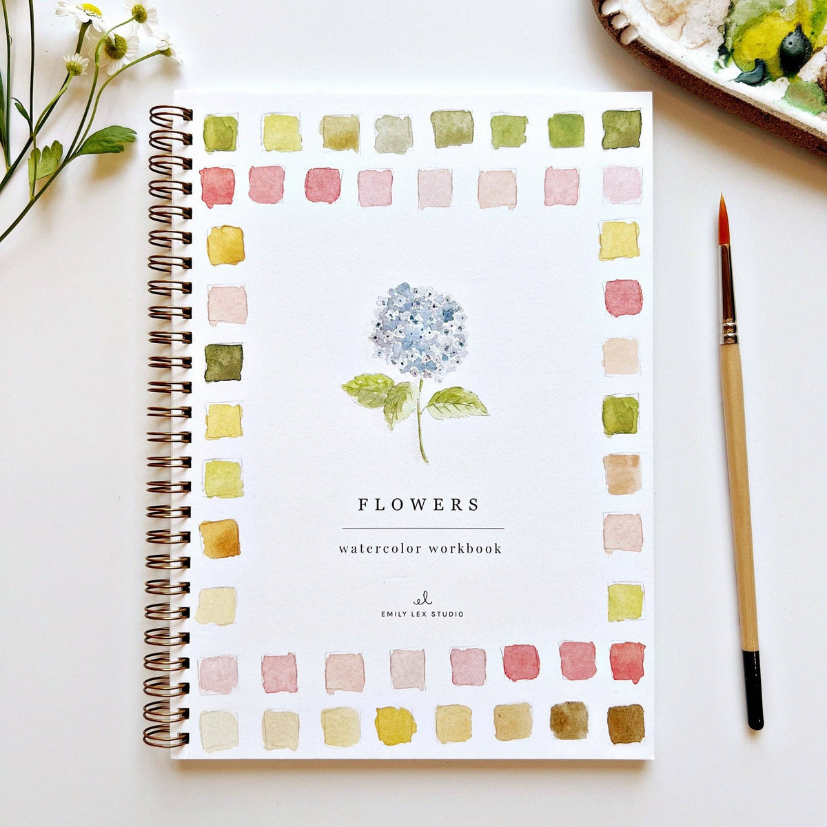 A spiral-bound watercolor workbook titled 'FLOWERS' with a central blue hydrangea illustration and a border of pastel watercolor swatches, shown on a white surface with a paintbrush and small flowers nearby.